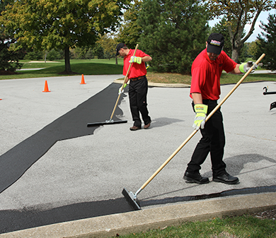 Sealcoating contractor applying white traffic marking paint in stripes in a freshly sealcoated parking lot.