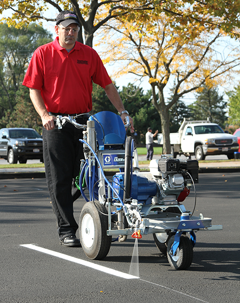 Sealcoating contractor applying white traffic marking paint in stripes in a freshly sealcoated parking lot.